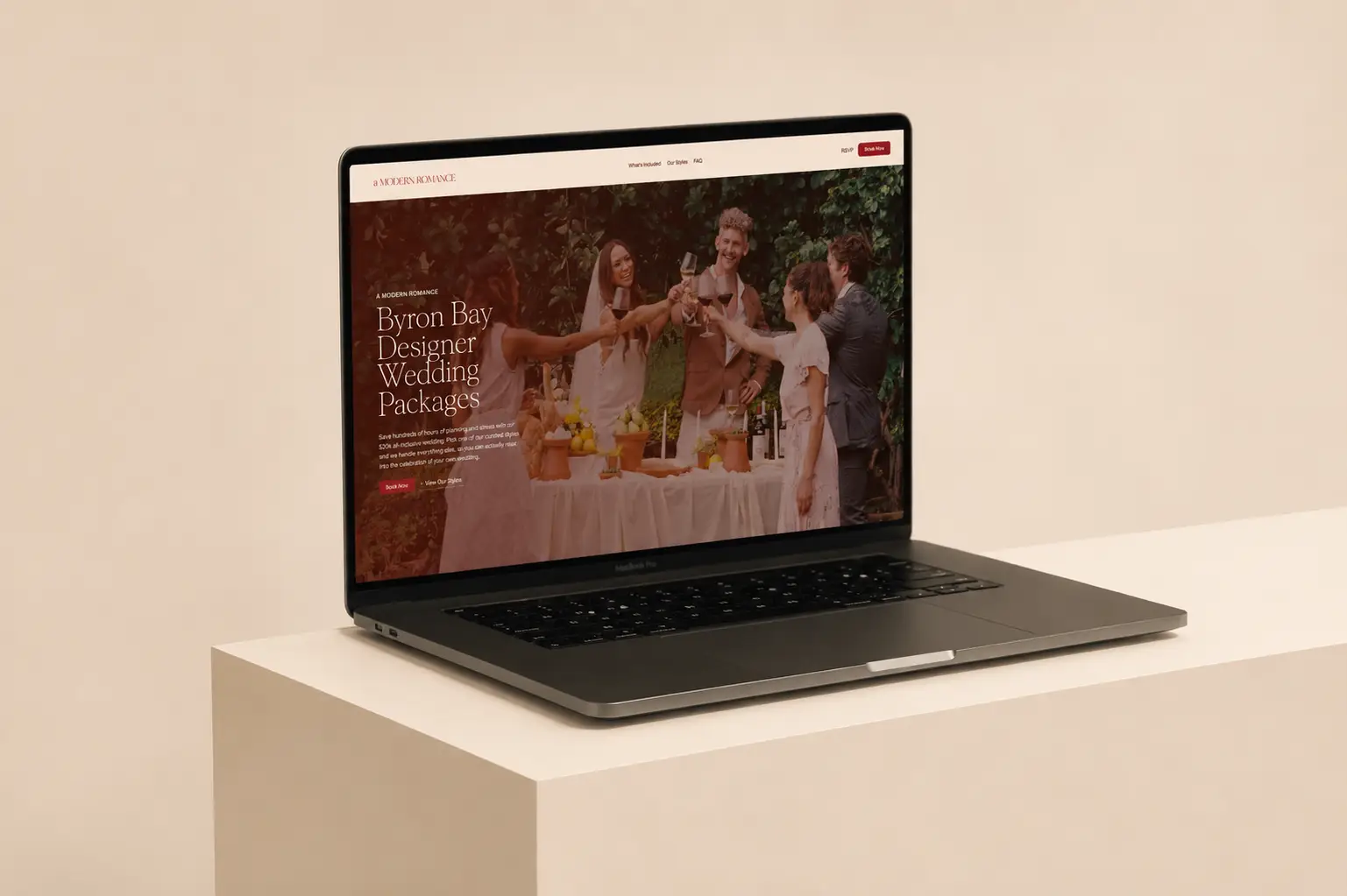 A MacBook Pro on a white plinth displays the A Modern Romance website homepage. A MacBook Pro on a white plinth displays the A Modern Romance website homepage. The hero shows a garden wedding toast scene with a warm red overlay, headlined "Byron Bay Designer Wedding Packages" with Book Now and View Our Styles CTAs.